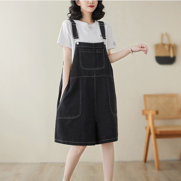 Person wearing a black denim pinafore dress with a white t-shirt underneath, standing in a room with wooden flooring and a chair.