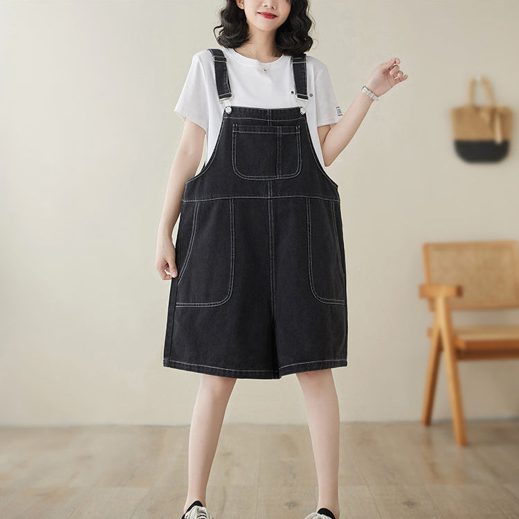 Woman wearing a black denim pinafore dress with a white shirt underneath, standing in a room with wooden flooring and a chair.