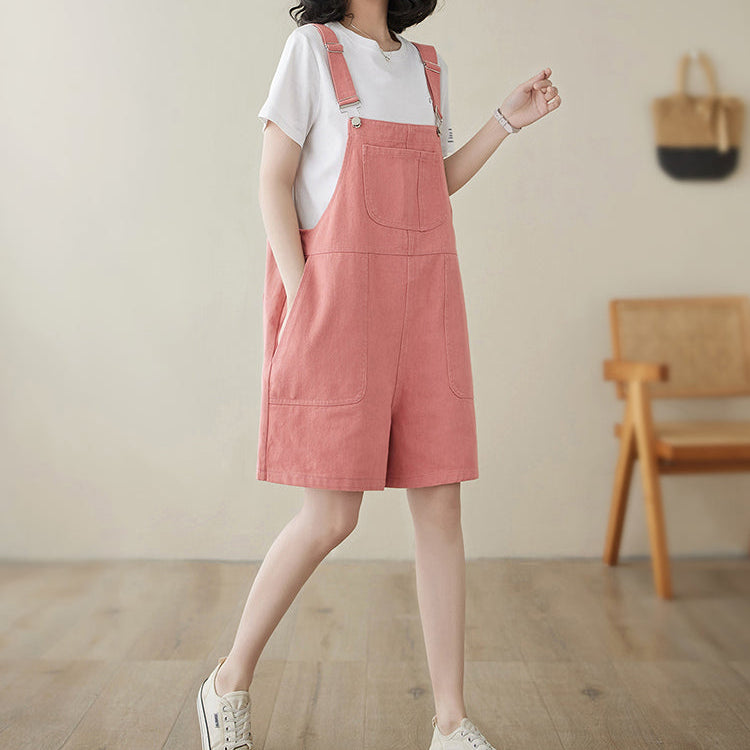 Woman wearing a pink denim pinafore dress with a white shirt underneath, standing in a room with wooden flooring and a chair.