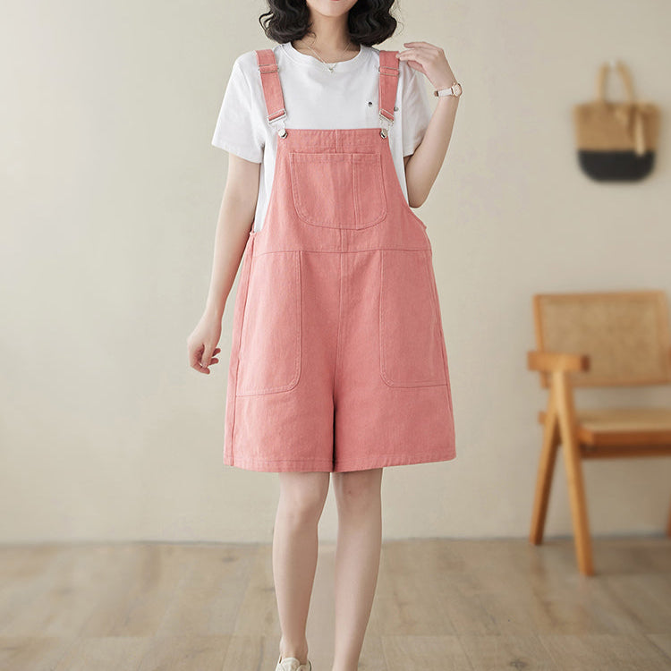 Woman wearing a pink denim overall dress with a white shirt underneath, standing in a room with wooden flooring and a chair.