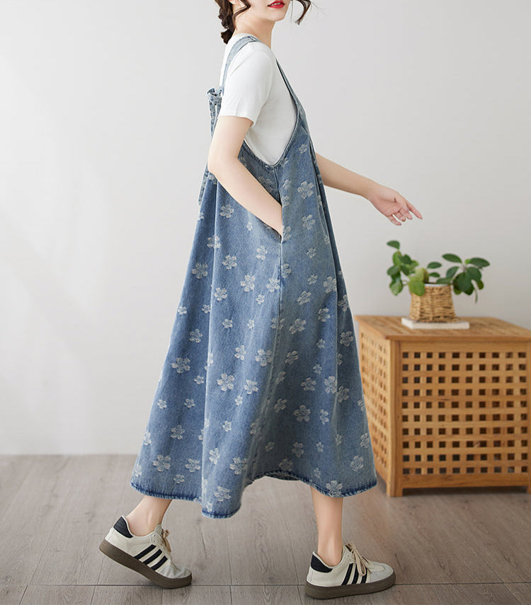 Woman wearing a blue floral overalls dress in a room with a wooden side table and plant.