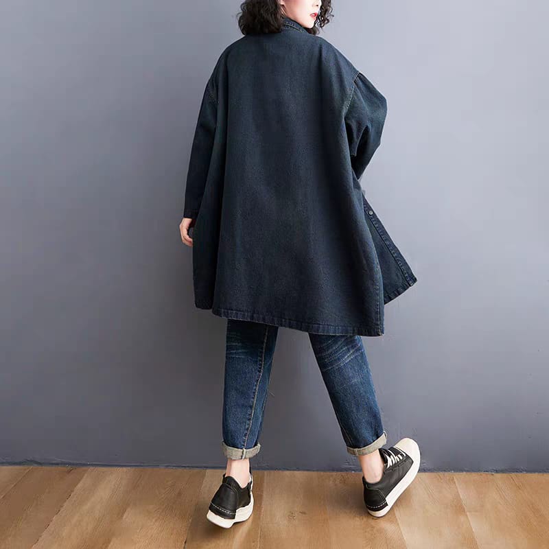 Person wearing a dark blue coat and jeans standing against a gray wall.