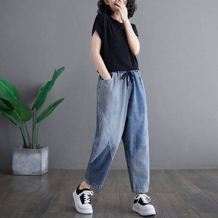 Person wearing a black top and light blue pants standing against a gray wall with a plant in the corner.