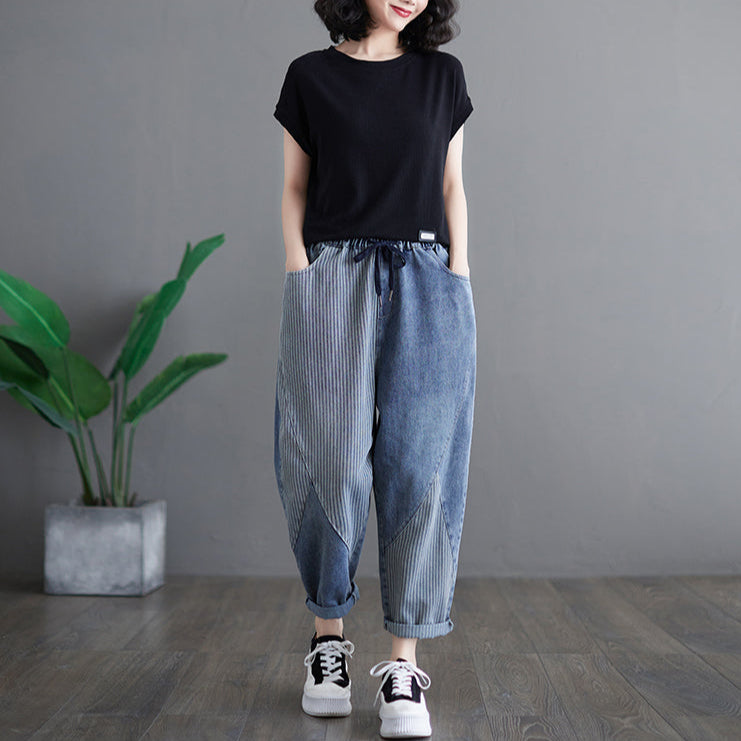 Person wearing a black t-shirt and blue striped pants standing in a room with a plant and gray wall.