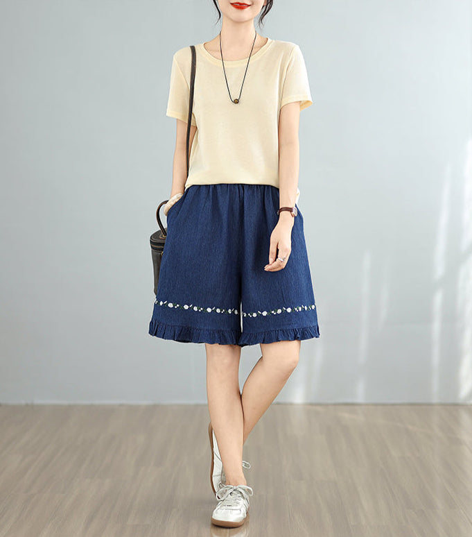 Woman wearing a beige top and blue skirt with white embroidery on a plain background
