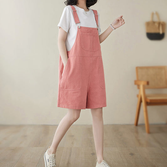 Woman wearing a pink denim pinafore dress with a white shirt underneath, standing in a room with wooden flooring and a chair.