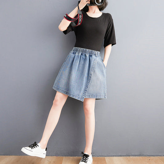 Person wearing a black t-shirt and denim skirt with white sneakers against a gray wall.