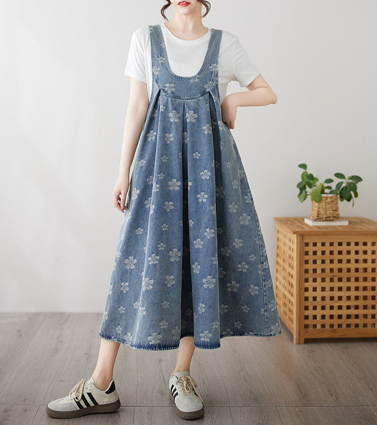 Person wearing a blue floral overalls dress with a white shirt underneath, standing in a room with a wooden side table and plant.