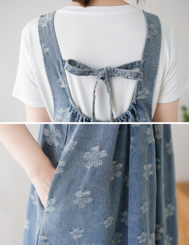 Denim pinafore dress with floral patterns worn over a white shirt on a neutral background.