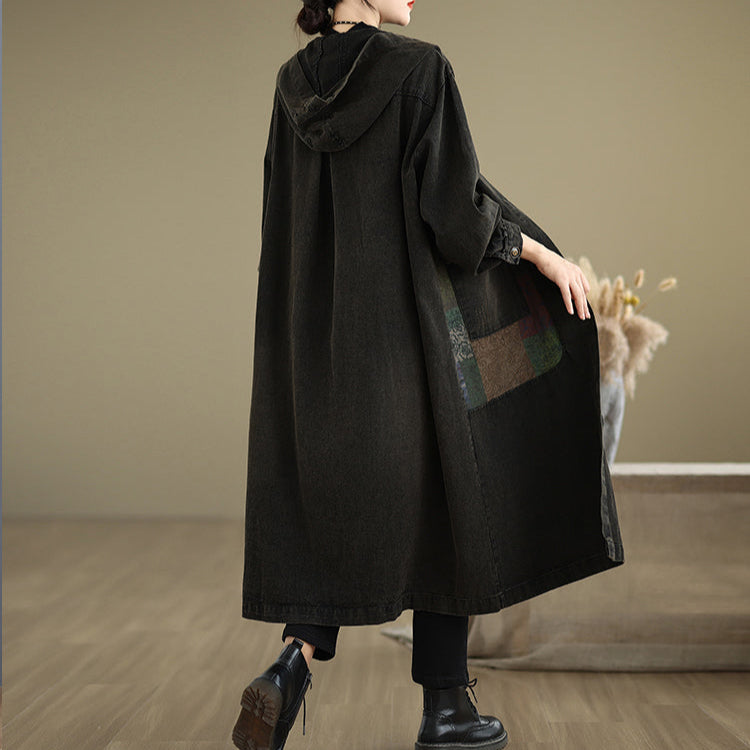 Person wearing a long black coat with a plaid patch in a minimalistic room.